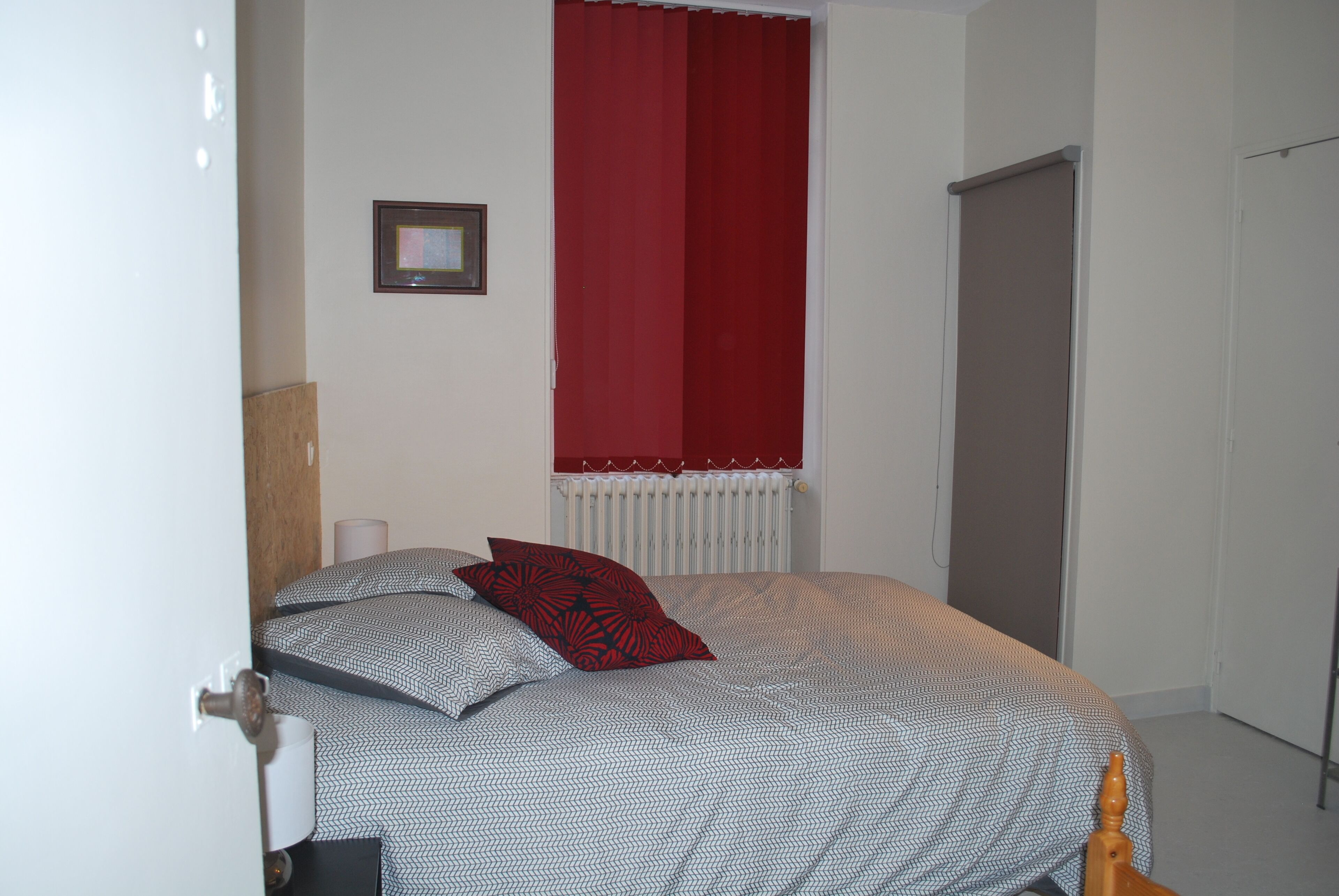 A bedroom with a bed, a window with red curtains, a radiator, and a door.