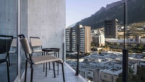 Un balcon agrémenté de fauteuils en osier et d’une petite table, d’où l’on peut admirer un paysage urbain avec des bâtiments modernes et une montagne en arrière-plan.
