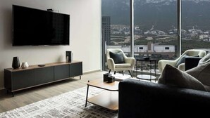 A modern living room with a large TV, a wooden coffee table, and a sofa. There are two chairs and a small table. The room has large windows with a mountain view. - Mountain View in Valle Poniente, Rooftop Pool, Gym (Santa Catarina)
