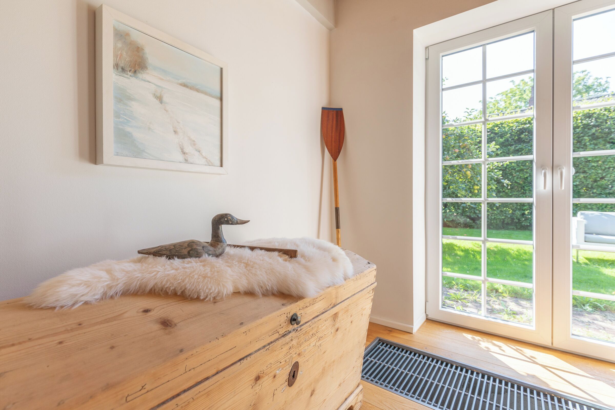 A wooden chest with a bird and a paddle leaning against a wall, a painting above it, and a view of greenery through a glass door.