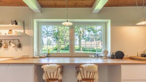 Eine Küche mit großem Fenster, Holzboden, weißen Schränken und zwei Stühlen mit weichen Sitzflächen.