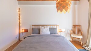 Ein Schlafzimmer mit einem Holz-Kopfteil, einem Bett mit grauer Bettwäsche, zwei Nachttischlampen, einer Pendelleuchte und einem Holzstuhl.
