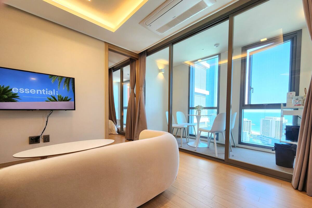 A modern hotel room with a large window, a flat-screen TV mounted on the wall, a white sofa, and a small table.