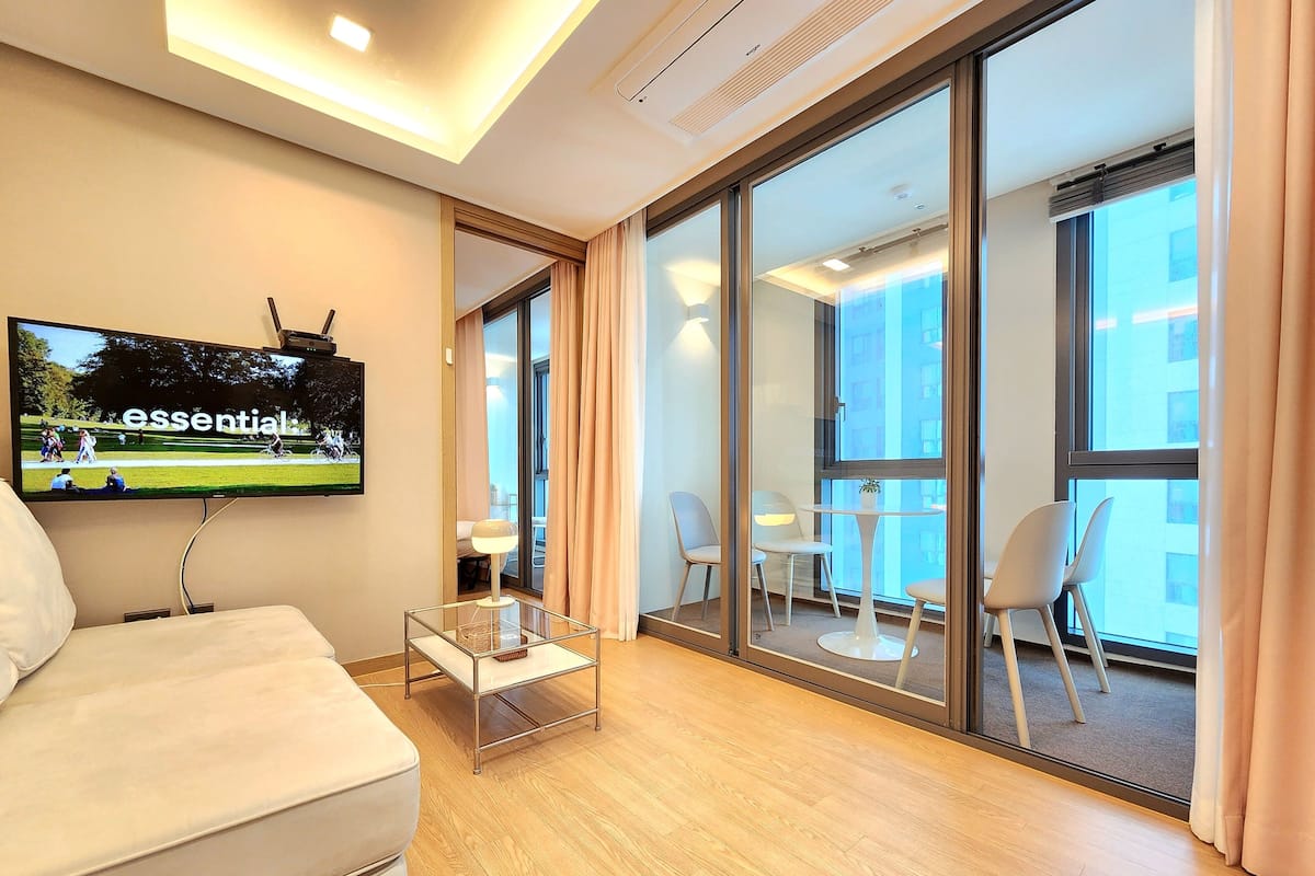 A modern hotel room with a large TV, a sofa, a glass table, and a balcony with chairs.