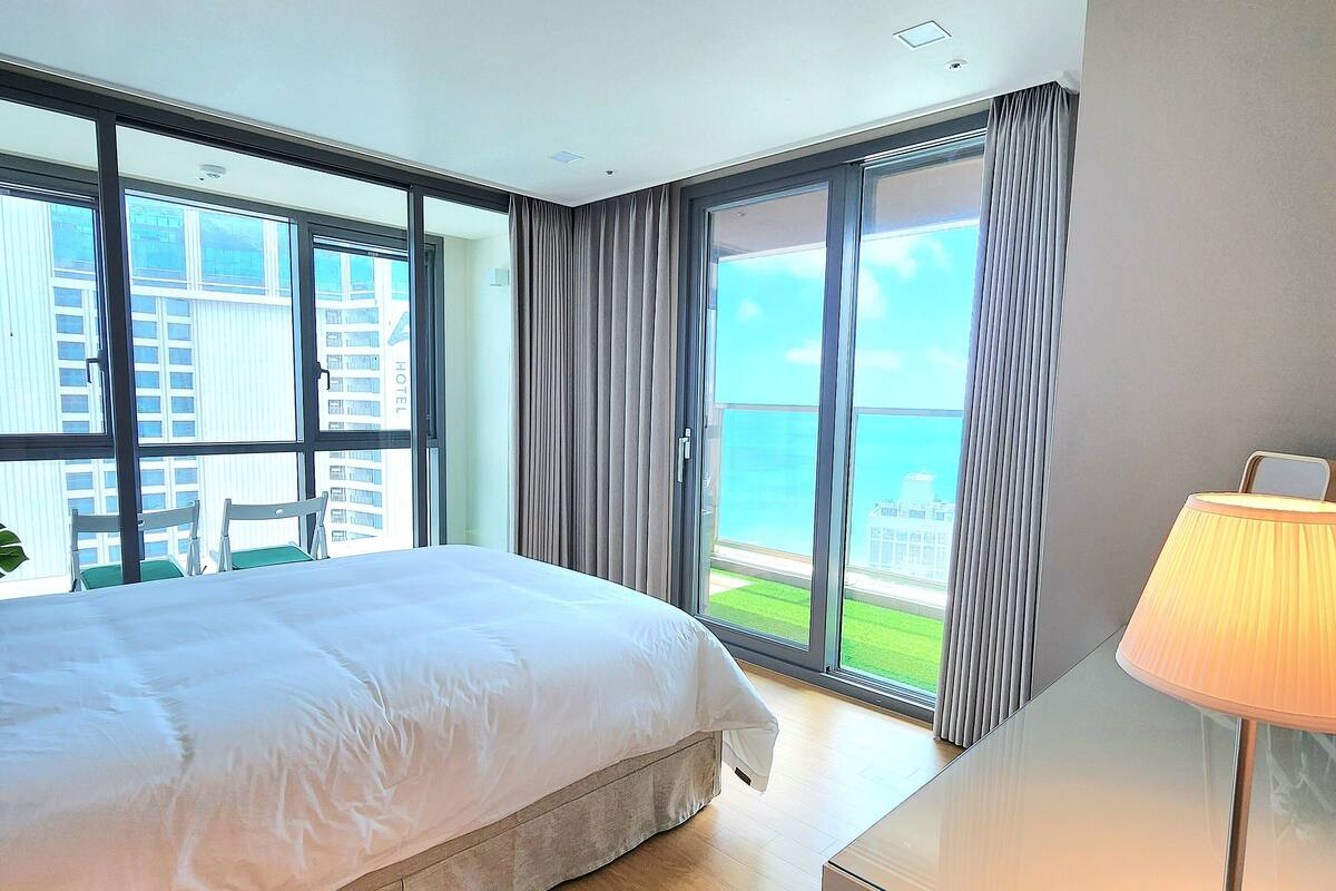 A hotel room with a large bed, a desk, and a view of the city through floor-to-ceiling windows.