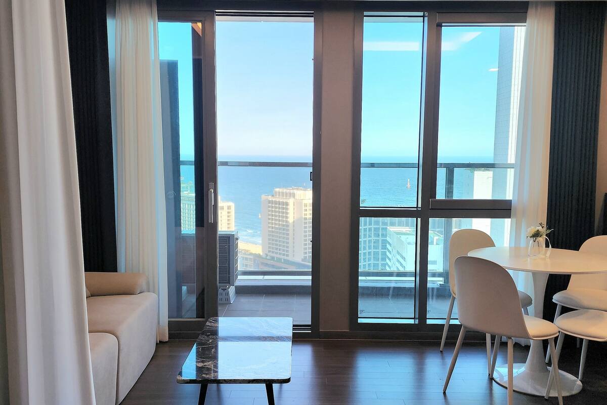 A modern hotel room with a large balcony, a dining area, and a view of the city and sea.