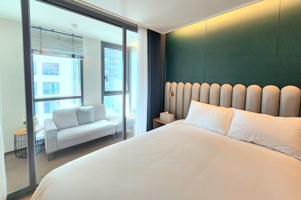 A modern hotel room with a large bed, a sofa, and a view of the city through floor-to-ceiling windows.