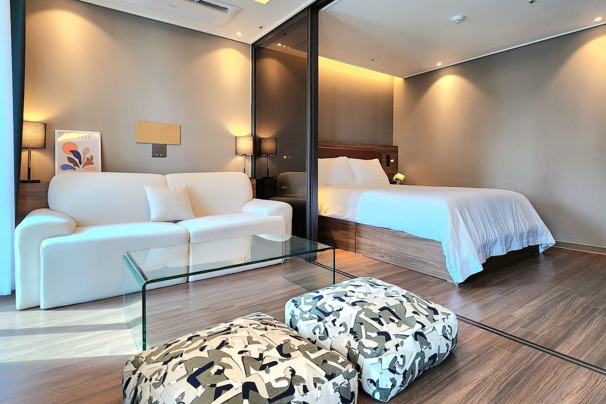 A modern hotel room with a white sofa, a glass coffee table, patterned ottomans, a bed with white linens, and a large mirror reflecting the room.