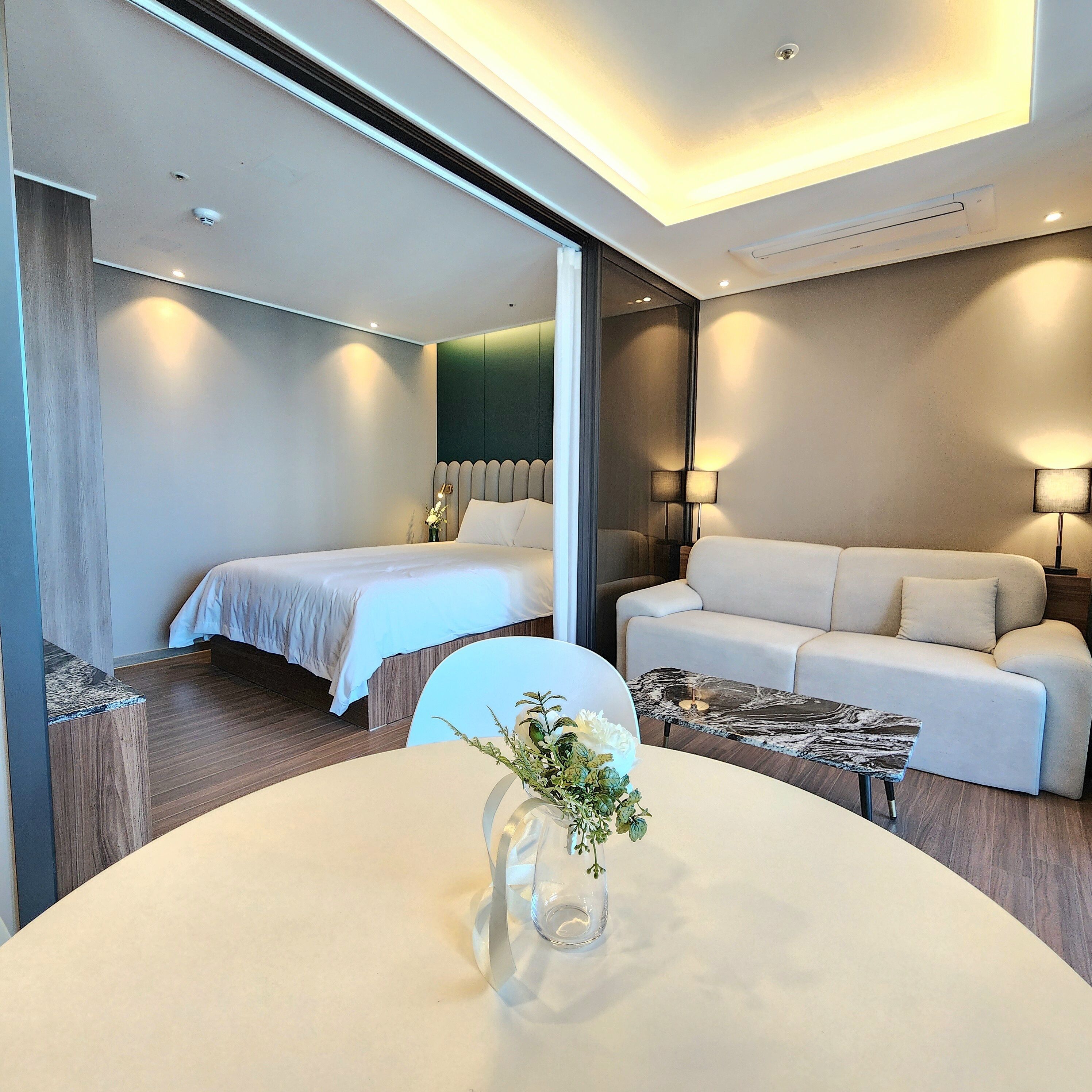 A modern hotel room with a bed, a sofa, a round table with a vase, and a mirror.
