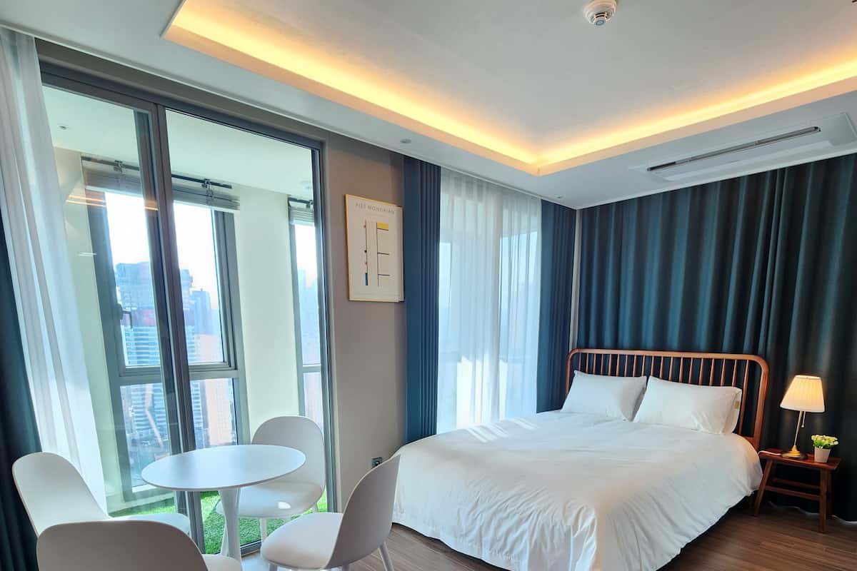 A hotel room with a bed, a round table with chairs, a lamp, and a view of the city through large windows.