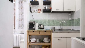 Una cocina compacta con armarios blancos, un lavamanos de acero inoxidable y una estantería de madera con vajilla.