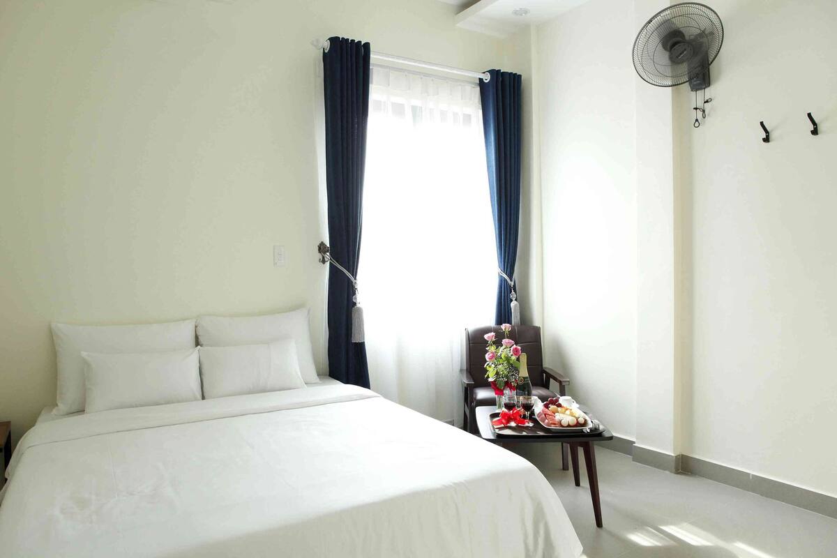 A hotel room with a bed, a chair, a table with a tray of food, and a wall-mounted fan.