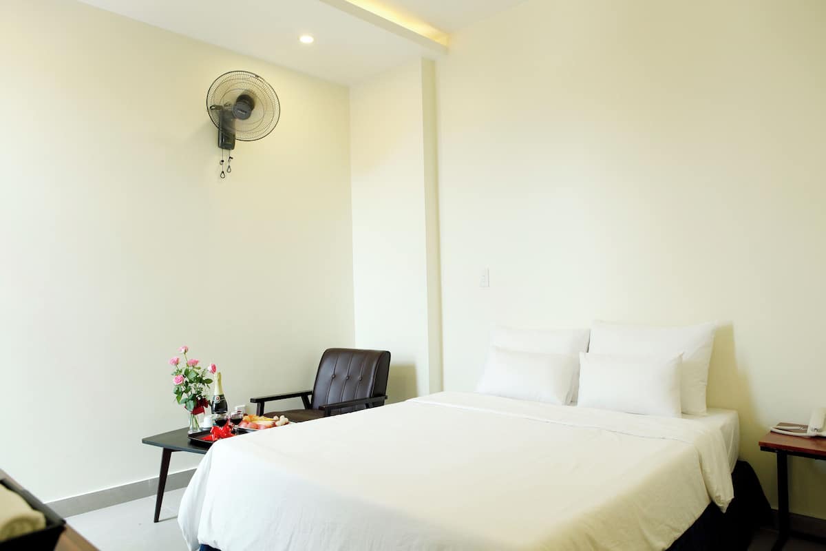 A hotel room with a bed, a chair, a small table with a vase of flowers, and a wall-mounted fan.