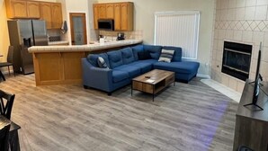 A modern living room with a blue sectional sofa, a wooden coffee table, and a kitchen area with wooden cabinets and a refrigerator.
