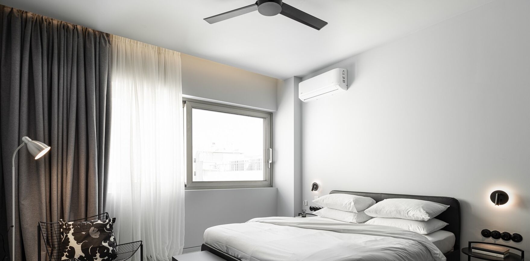 Ett modernt hotellrum med en säng, en takfläkt, ett fönster med gardiner och en väggmonterad luftkonditioneringsanläggning.