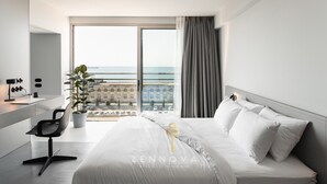 A modern hotel room with a large bed, a desk with a chair, a potted plant, and a view of the sea and buildings. - Nero Luxury Suites by Zennova (Thessaloniki)