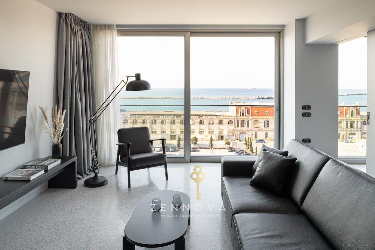 Una sala de estar moderna con un sofá, una butaca, una mesa de centro y un televisor. Se ve un ventanal grande con vistas a un edificio y al mar.