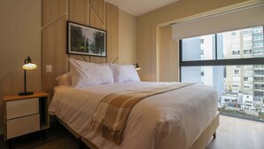 Een slaapkamer met een groot bed, een nachtkastje met een lamp, een ingelijst schilderij aan de muur en een raam met uitzicht over de stad.