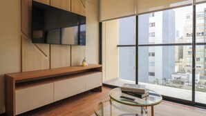 Een moderne woonkamer met een houten console, een flatscreen televisie aan de muur, een glazen salontafel en een groot raam met uitzicht op de stad.