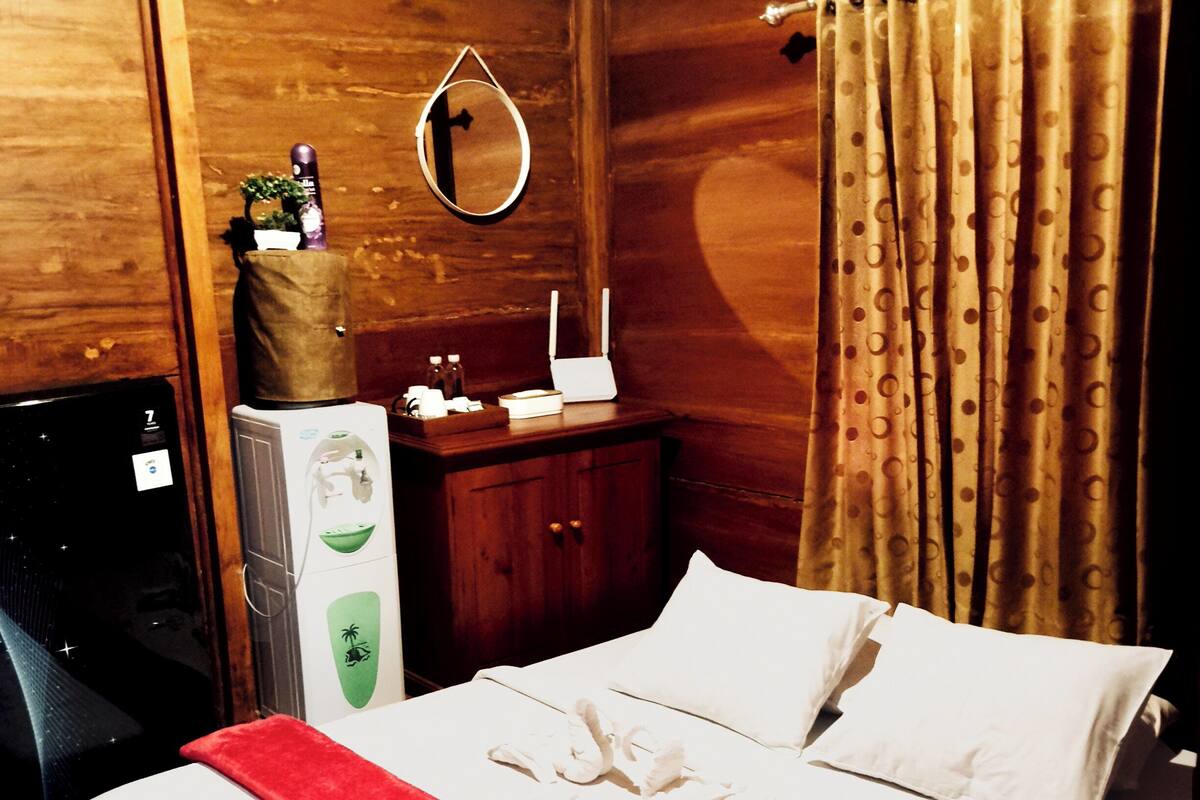 A hotel room with a bed, a wooden wardrobe, a desk with a computer, a small refrigerator, and a window with curtains.