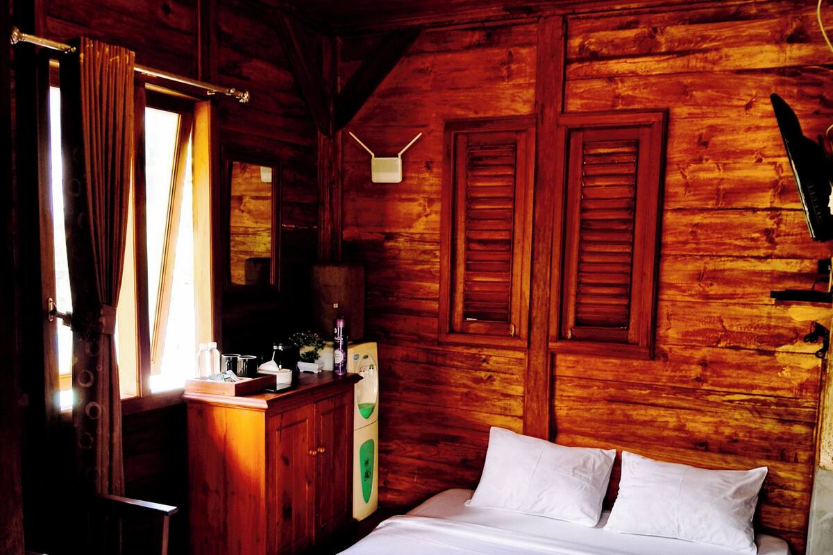 A wooden cabin room with a bed, a dresser, and a window with curtains.