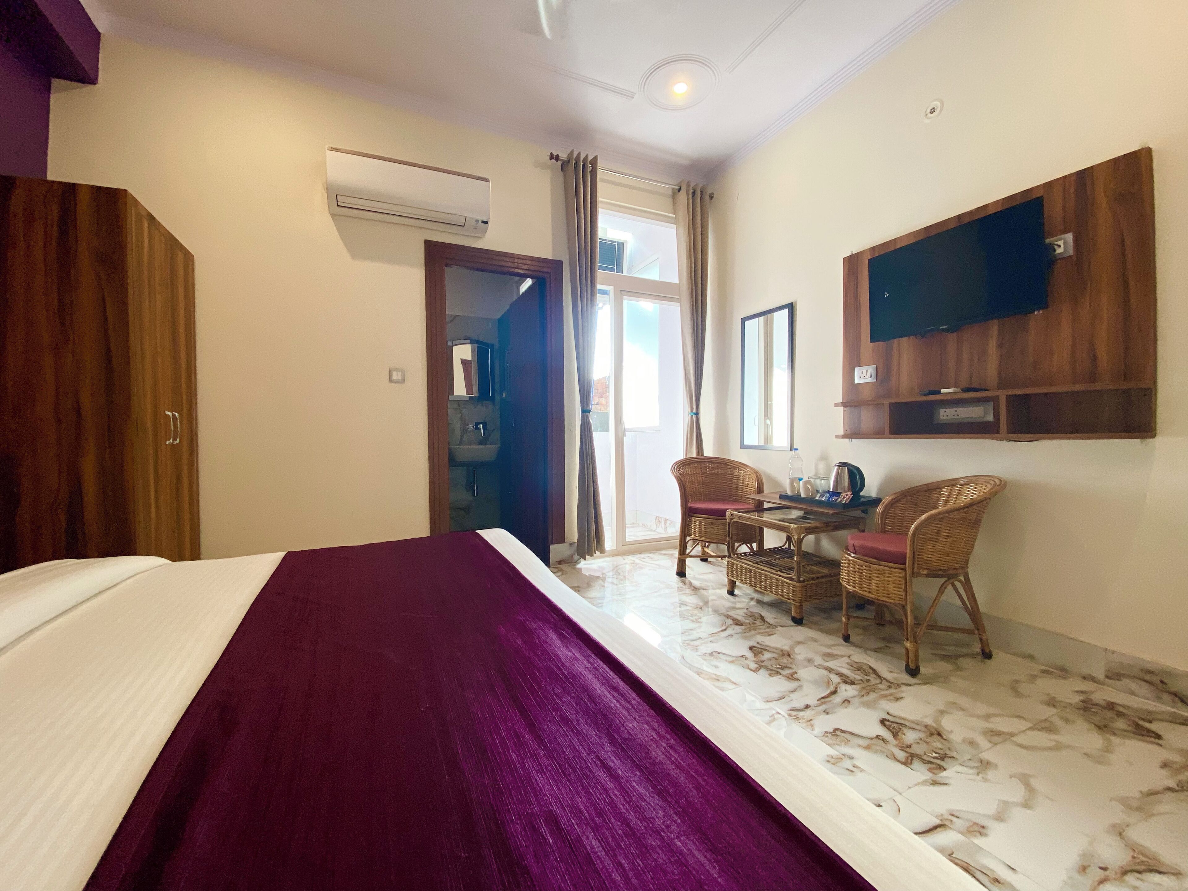 A hotel room with a bed, a desk with chairs, a TV, and an air conditioning unit.