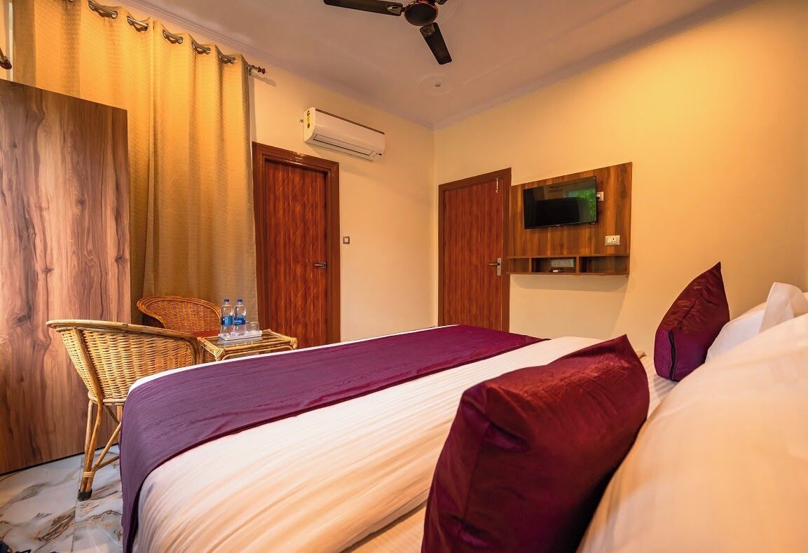A hotel room with a bed, a television mounted on the wall, a chair, and an air conditioning unit.