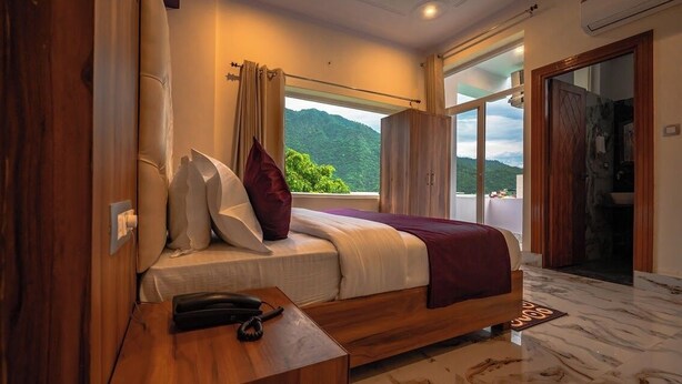 A bedroom with a bed, a bedside table with a phone, a window with curtains, and a view of greenery. - Hotel Rishikesh Grand by Kool-stays (Rishikesh)