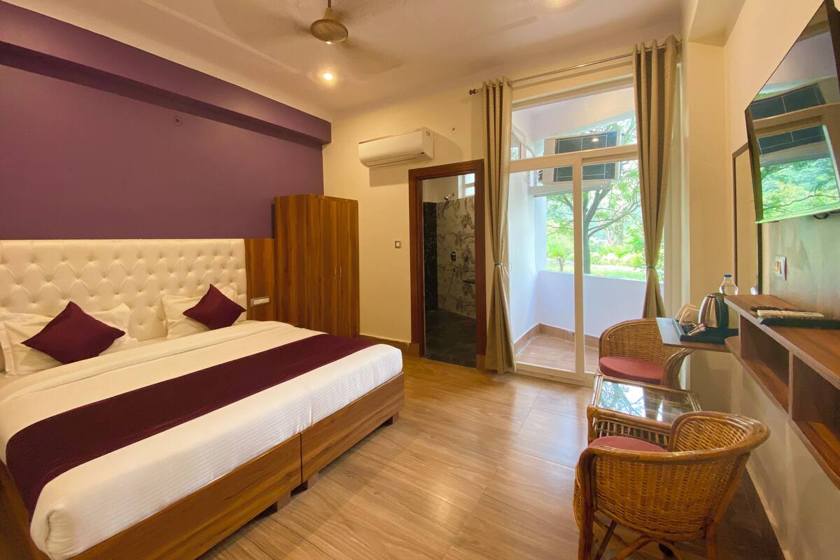 Una habitación de hotel con una cama grande, un armario de madera, una mesita con sillas, televisión y un ventanal corredero que da a un balcón.