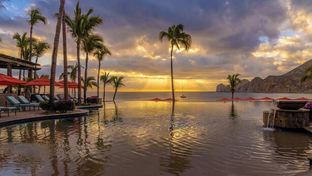 Piscina de un resort con palmeras y sombrillas rojas, con vista al mar tranquilo al atardecer.