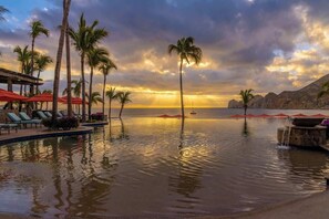 A resort pool with palm trees and red umbrellas, overlooking a calm sea at sunset.
