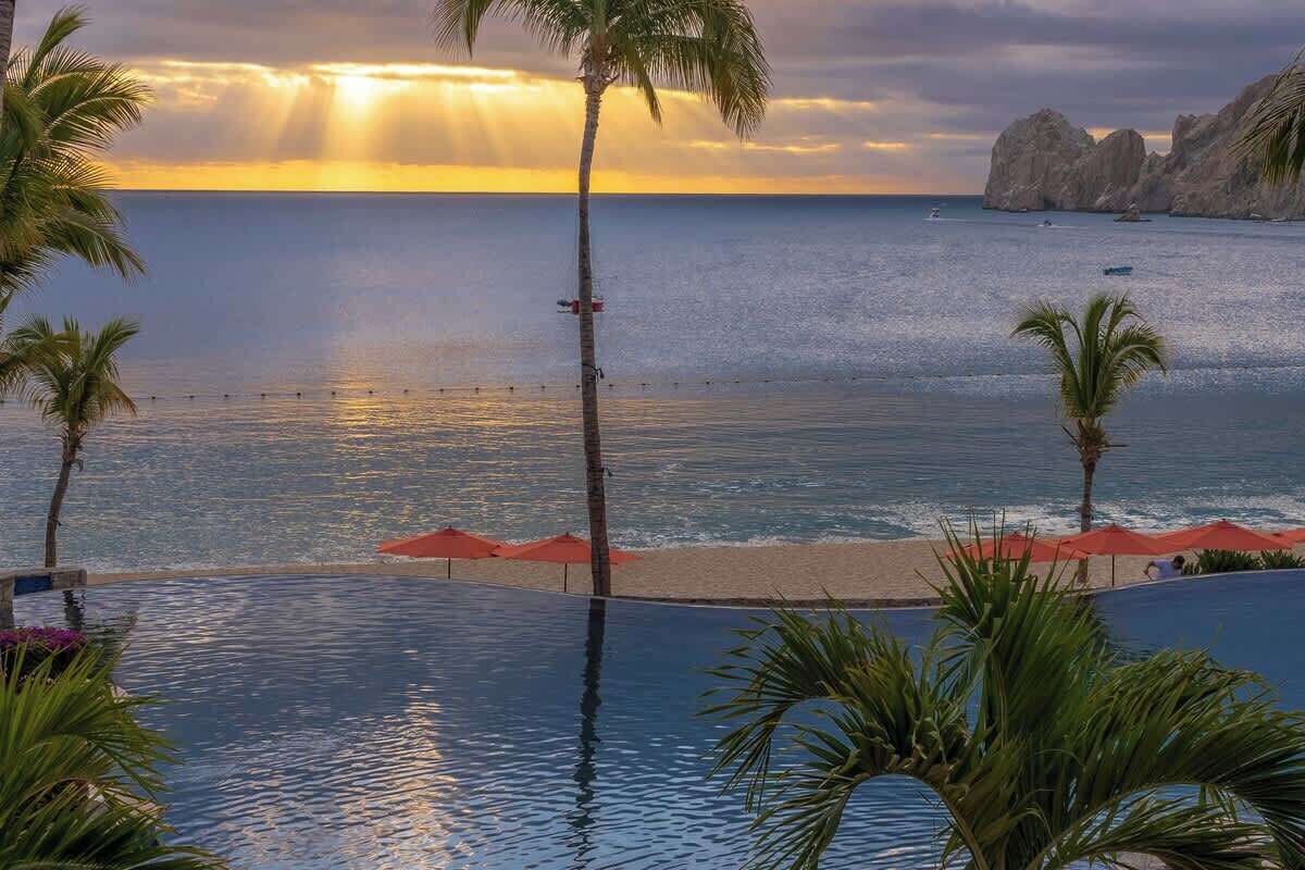 A poolside view with a sunset over the ocean, palm trees, and a rocky coastline.