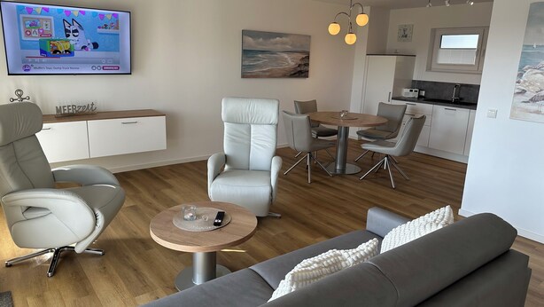 Ett modernt vardagsrum med en grå soffa, ett runt soffbord och en matplats med ett litet bord och stolar. En tv är monterad på väggen, och ovanför matbordet hänger en tavla.