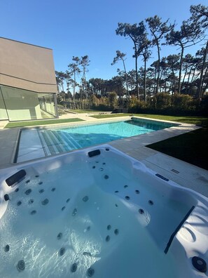 Une piscine avec un jacuzzi, entourée d’un jardin aménagé et de grands arbres.