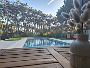 A swimming pool with a wooden deck in the foreground, surrounded by trees and a clear sky.