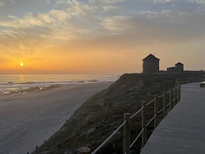 A coastal scene with a wooden boardwalk, a beach, and two stone structures.