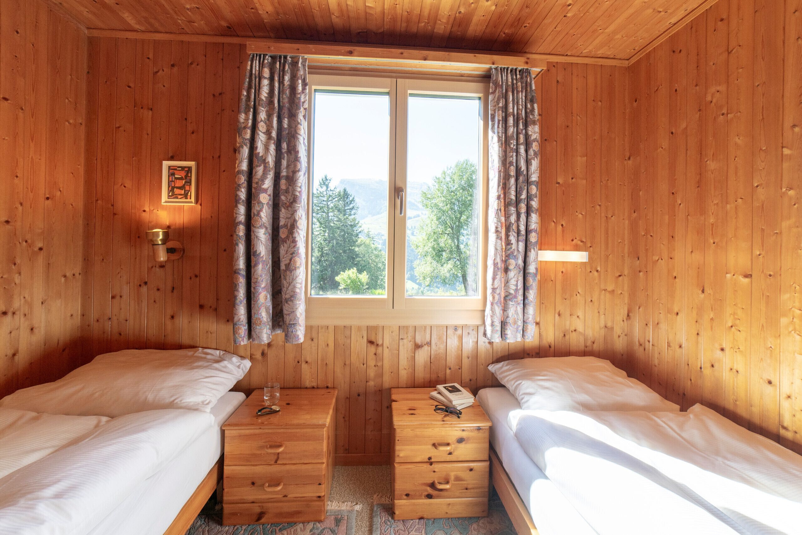 Ein Zimmer in einer Holzhütte mit zwei Betten, einem Fenster mit Vorhängen, einem kleinen Tisch mit einer Lampe und einem Bild an der Wand.