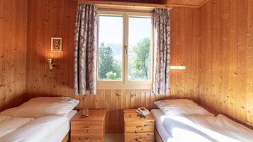 Ein Zimmer in einer Holzhütte mit zwei Betten, einem Fenster mit Vorhängen, einem kleinen Tisch mit einer Lampe und einem Bild an der Wand.