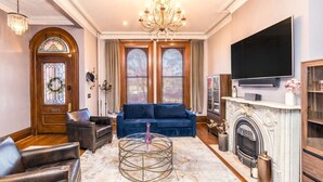 Een woonkamer met een blauwe bank, leren fauteuils, een kroonluchter, een open haard en een flatscreen televisie.