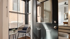 Una habitación con una puerta negra, una silla de mimbre con cojín, una pequeña mesa redonda con una planta y vistas a un edificio de ladrillo a través de grandes ventanales.
