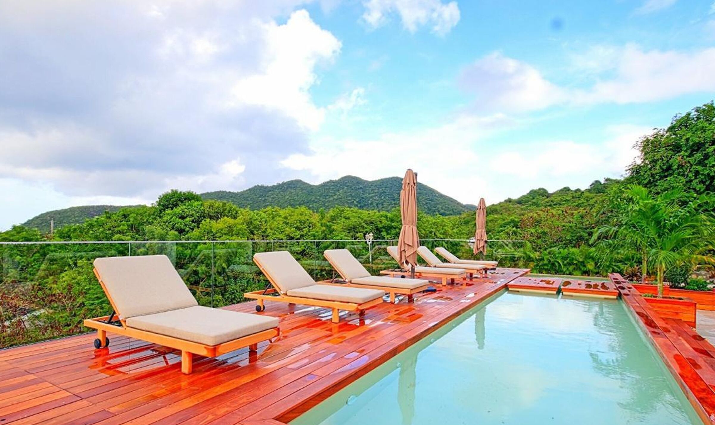 A poolside area with lounge chairs and umbrellas, surrounded by lush greenery and mountains.