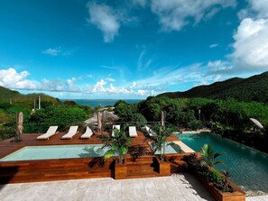 A pool area with lounge chairs and wooden decking, surrounded by lush greenery and a view of the sea. - Apartment with Private Pool and Garden View (Saint-Martin)