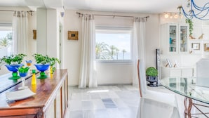 A modern dining area with a glass table, white chairs, and a view of palm trees through a window.