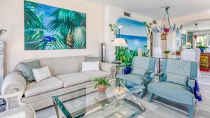 A living room with a sofa, a glass coffee table, and a large tropical-themed painting.