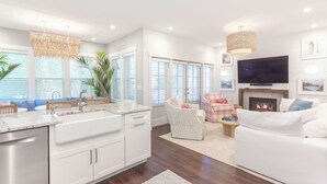 A modern kitchen with white cabinets and a large island, a living room with a white sofa and a fireplace, and a dining area with a checkered tablecloth. - Designer Downtown Home, Assn Pool, Walk To Beach (New Buffalo)