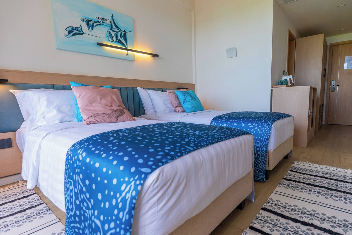 A hotel room with two beds, a wooden headboard, a wall-mounted light, and a painting of a fish.