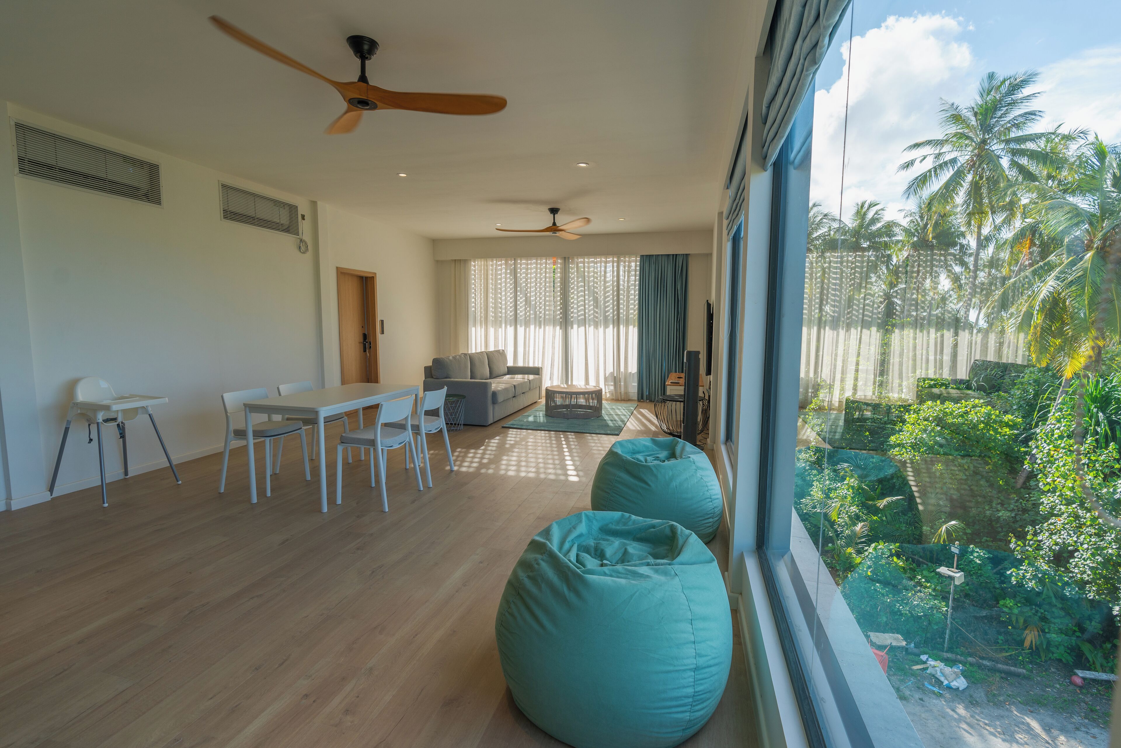 Ein geräumiger Wohnbereich mit Esstisch, Stühlen, einer Couch und zwei Sitzsäcken. Es gibt Deckenventilatoren, ein großes Fenster mit Blick auf einen Garten und eine Tür zu einem anderen Raum.