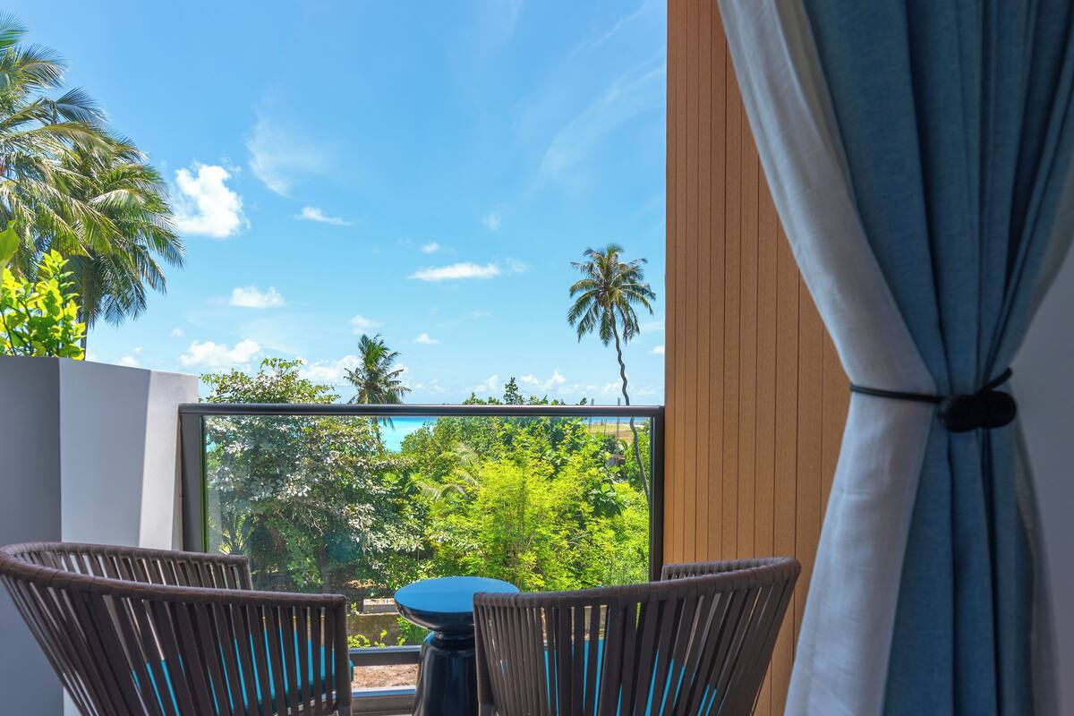 A balcony with two wicker chairs and a small table, overlooking a lush green landscape and palm trees under a clear blue sky.