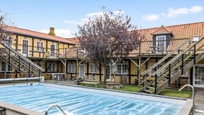 A swimming pool with wooden deck and stairs leading to a building with yellow walls and red-tiled roofs.