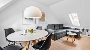 A modern, well-lit room with a round dining table, black chairs, a gray sofa, and a small side table. There are framed pictures on the wall and a skylight above the sofa.